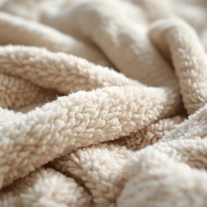 May include: Close-up of a soft, beige, plush fabric. The material has a thick, textured surface with a high pile, creating a warm and inviting appearance. The folds and curves of the fabric add depth and dimension.
