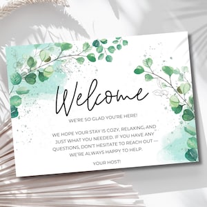 Puede incluir: Una tarjeta de bienvenida blanca con ilustraciones de eucalipto y la palabra "Welcome" en escritura cursiva. La tarjeta incluye el texto "We're so glad you're here!" y un mensaje sobre la estancia. Acentos de acuarela verdes y azules decoran la tarjeta.