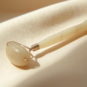 May include: A cream-colored facial roller with a rounded head and handle. The roller is made of a smooth, possibly jade-like material, and is attached to a silver-colored metal frame. The roller is resting on a cream-colored fabric.
