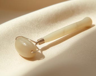 May include: A cream-colored facial roller with a rounded head and handle. The roller is made of a smooth, possibly jade-like material, and is attached to a silver-colored metal frame. The roller is resting on a cream-colored fabric.