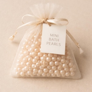 May include: A small, sheer, ivory-colored bag filled with mini bath pearls. The bag is tied with a matching ribbon and a tag that reads "MINI BATH PEARLS". The pearls are a creamy, off-white color.