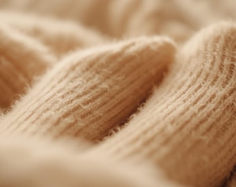May include: A close-up view of a beige knitted fabric featuring a distinct ribbed texture. The material displays a soft, fuzzy surface with vertical lines created by the knit pattern, showing the intricate loops and fibers of the light tan yarn.