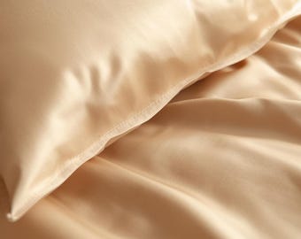 May include: Close-up of a beige satin pillow and bedding. The fabric has a smooth, lustrous appearance with subtle folds and shadows, suggesting a luxurious texture. The stitching on the pillow's edge is visible.