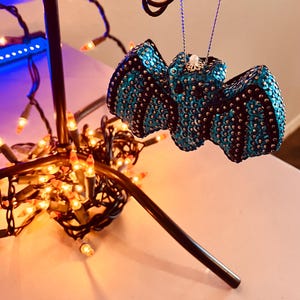 May include: A blue and black sparkly bat ornament hanging from a string of lights. The bat is covered in sequins and has a shiny finish.