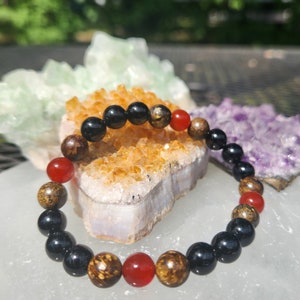 May include: A black, brown, and red beaded bracelet with a mix of round and oval beads. The bracelet is on a white surface with a yellow and purple crystal in the background.