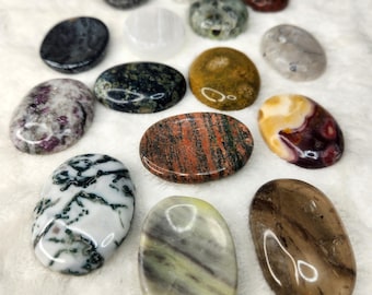 May include: Assortment of oval-shaped polished stones in various colors and patterns, including red, brown, white, green, and yellow. The stones are arranged on a white fuzzy surface.