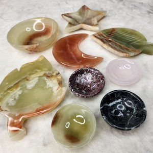 May include: Assortment of nine small, decorative bowls made of various types of stone. The bowls are in different shapes, including a star, a crescent moon, a leaf, and a circle. The bowls are in various colors, including green, brown, pink, and black.