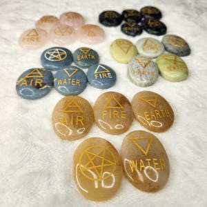 May include: Set of 12 smooth, oval-shaped stones with gold painted symbols and text. The symbols represent the four elements: air, fire, water, and earth. The stones are a light brown color.