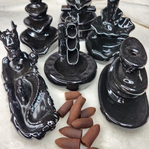May include: A collection of black ceramic incense burners in various shapes, including stacked designs, a mermaid, and a traffic light. Brown incense cones are also present, offering a complete set for aromatherapy or spiritual practices.