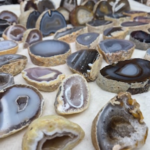 May include: A collection of numerous agate geode slices featuring varied concentric banding patterns in shades of gray, brown, white, and black. Each piece displays a rough, textured outer edge and a polished, smooth interior surface with crystalline centers.