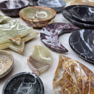 May include: A collection of decorative stone dishes and trays in various shapes and colors. The assortment includes bowls, star-shaped trays, leaf-shaped trays, and crescent-shaped trays. The colors range from green, brown, and white to black and gray, with unique veining patterns.