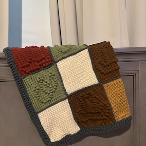 Può includere: Una coperta quadrata all'uncinetto fatta a mano con un design patchwork. La coperta presenta quadrati nei toni del rosso, verde, marrone, crema e oro, con motivi a forma di cuore. Il bordo è grigio scuro.