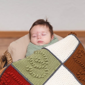 May include: A baby sleeping in a wicker basket, covered by a handmade crochet blanket featuring square patches in shades of cream, olive green, rust red, and brown, each decorated with a raised textured heart pattern and dark gray borders.