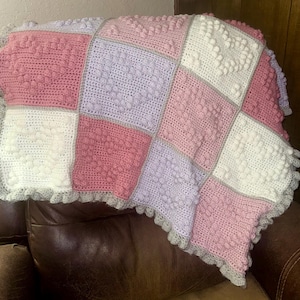 May include: A handmade crochet blanket in a patchwork design. The blanket features squares of pink, white, and lavender, with a gray border and ruffled edge. The blanket is resting on a brown leather chair.