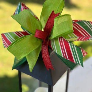 May include: A black lantern with a green and red striped ribbon bow and a red ribbon with glitter. The lantern has a glass pane and a white candle inside.