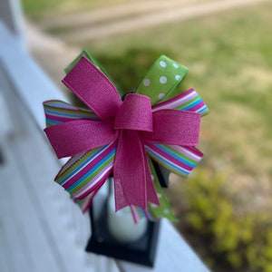 May include: A pink, green, and white ribbon bow tied on a black lantern. The bow is made of multiple ribbons with different patterns, including polka dots and stripes.