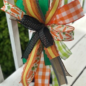 May include: A decorative bow featuring a variety of ribbons in spring colors. The bow includes ribbons with carrot, gingham, and polka dot patterns. The color palette includes orange, green, yellow, black, and white. The bow is likely for seasonal or holiday decor.