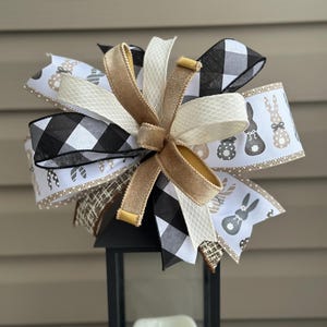 May include: A decorative bow featuring a mix of ribbons in black and white buffalo check, cream, and tan. Some ribbons have bunny and egg patterns. The bow is centered on a black lantern, creating a festive Easter decoration.