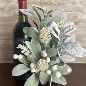 May include: A bottle of red wine with a decorative floral arrangement. The arrangement features green and gray leaves, white berries, and small beige pinecones. The wine bottle has a dark label and a red top.