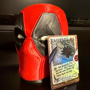 May include: A 3D-printed Deadpool mask in red and black, with white eye openings. A card with the text "Blacker Lotus" and instructions is propped up in front of the mask. The card has a Deadpool logo.