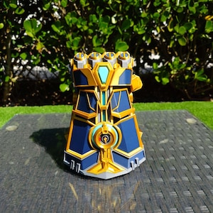 Könnte beinhalten: Ein detaillierter, gold- und blaufarbener Handschuh mit geometrischem Design. Der Handschuh hat einen zentralen türkis farbigen Edelstein und vier goldfarbene kreisförmige Akzente auf den Knöcheln. Die Basis ist silbergrau.