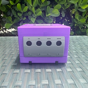 May include: A purple cube-shaped gaming console replica featuring a gray front panel with four circular controller ports and two memory card slots labeled SLOT-A and SLOT-B. The top surface includes a circular disc drive cover.