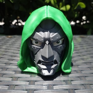Op de afbeelding: Een 3D-geprint borstbeeld van een personage met een felgroene capuchon en een metallic grijs masker. Het masker heeft een stijf, hoekig ontwerp met zichtbare klinknagels, een puntige neus en een strenge uitdrukking met zichtbare ogen.