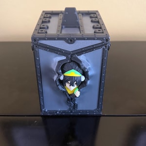 Op de afbeelding: Een grijze doosvormige container met een handvat aan de bovenkant en een karakterfiguur dat uit de voorkant komt. Het beeldje heeft een geel shirt, een groene hoofdband en zwart haar. De doos heeft geklonken metalen accenten.