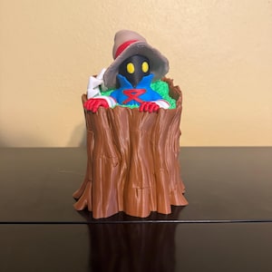 May include: A whimsical figurine of a character in a tree stump. The character has a black face, blue robe, red gloves, and a brown hat. The tree stump is brown with detailed bark texture, and the character is surrounded by green foliage.