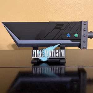 May include: A black and grey Buster Sword replica from Final Fantasy VII, displayed on a stand. The sword has a detailed design with blue and green accents. The Final Fantasy VII logo is visible on the stand.