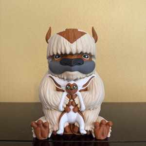 Op de afbeelding: Een beeldje van Appa, een fictieve vliegende bizon uit de animatieserie Avatar: The Last Airbender. Het beeldje is voornamelijk crèmekleurig met bruine accenten, waaronder de hoorns, de pijlvormige markering en de voeten van de bizon. Een kleiner figuur van Momo wordt in Appa's armen gehouden.