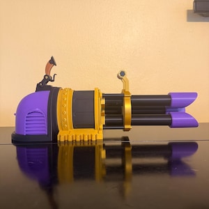 Puede incluir: Una pistola de juguete con una combinación de colores púrpura y dorado. La pistola presenta un diseño de cañón giratorio con múltiples cañones negros. La parte delantera y trasera de la pistola son púrpuras, y la sección media es dorada.