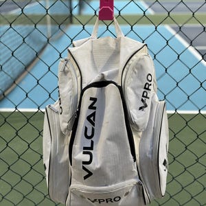 May include: A white and gray sports bag with black accents and the word "VPRO" and "VULCAN" printed on it. The bag is hanging on a pink hook attached to a chain link fence.