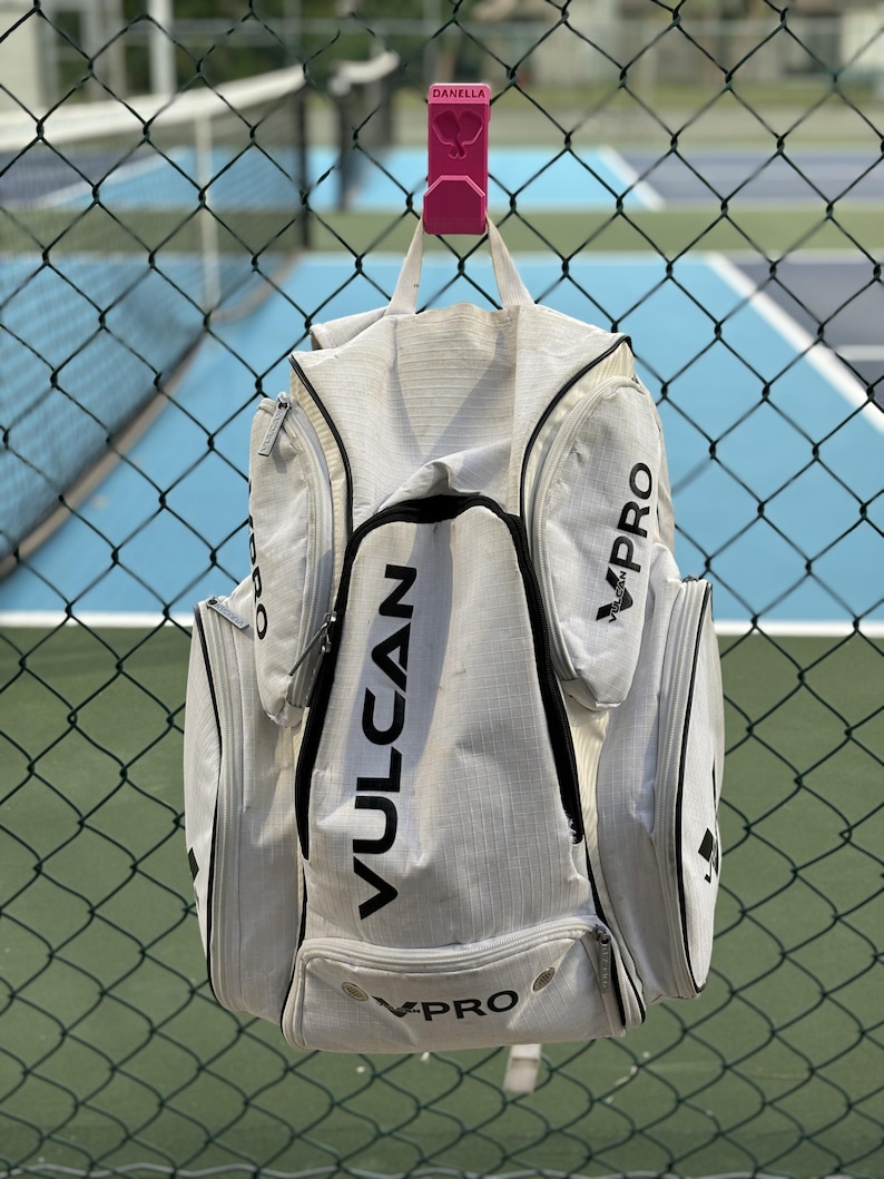 May include: A white and gray sports bag with black accents and the word "VPRO" and "VULCAN" printed on it. The bag is hanging on a pink hook attached to a chain link fence.
