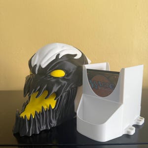 May include: A black, white, and yellow card holder shaped like a monster's head, with a separate white card holder. The monster head has a wide open mouth and yellow eyes. The white card holder has the words "MAGIC The Gathering" on the side.