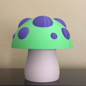 Puede incluir: Un objeto decorativo con forma de hongo. El sombrero es de color verde brillante con lunares morados, y el tallo es blanco. Esta pieza peculiar agregará un toque de fantasía a cualquier espacio.