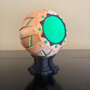 May include: A decorative sphere with a textured, segmented design in shades of tan, green, and black. The sphere has a bright green center and is mounted on a black pedestal. The overall design is geometric and modern.