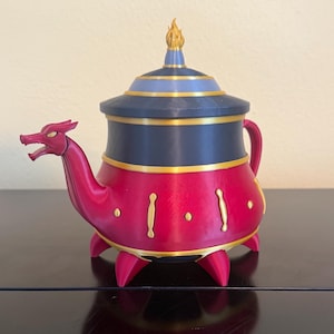 Op de afbeelding: Een decoratieve theepot met een draakvormige tuit en handvat. De theepot is voornamelijk rood met gouden accenten en een donkerblauwe deksel. De deksel is bekroond met een gouden, vlamvormige bekroning. Uniek, fantasie-geïnspireerd ontwerp.