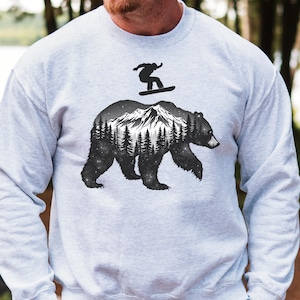 May include: A light gray sweatshirt featuring a black and white graphic of a bear with a mountain and forest scene inside its silhouette. A snowboarder is depicted above the bear. The sweatshirt is made of a soft material.