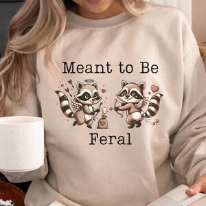 May include: A beige sweatshirt featuring a graphic of two cartoon raccoons with angel wings. The raccoons are holding a heart and an arrow. The text "Meant to Be Feral" is printed on the sweatshirt.