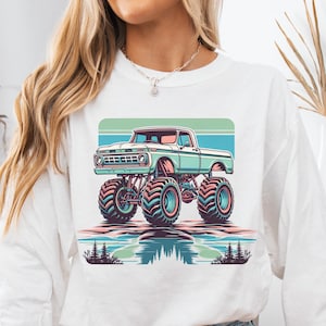 May include: A white long-sleeved sweatshirt with a vintage-style graphic of a teal green monster truck with large tires. The truck is driving through a landscape with a blue, green, and pink sunset in the background.