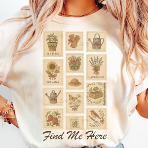 May include: Ivory-colored t-shirt featuring a collection of postage stamp-style illustrations of gardening elements. The stamps depict herbs, sunflowers, lavender, watering cans, and other garden-related items. The text "Find Me Here" is printed at the bottom.
