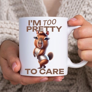 May include: White ceramic mug with a brown capybara wearing a white swimsuit and eyelashes. The mug has the text "I'm Too Pretty To Care" in brown letters.
