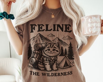 May include: A brown t-shirt with a black graphic of a cat wearing a backpack and standing in front of a tent. The text "Feline the Wilderness" is printed below the cat.