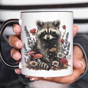 Puede incluir: Taza de cer&aacute;mica blanca con asa y borde negros. La taza presenta una ilustraci&oacute;n detallada de un mapache rodeado de flores, setas y follaje. El mapache tiene pelaje gris y negro, y las flores son rojas, blancas y amarillas.
