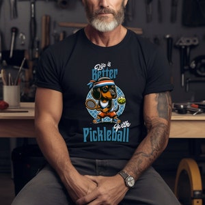 K&ouml;nnte beinhalten: Ein schwarzes T-Shirt mit einem Cartoon-Dackel, der Pickleball spielt. Der Text auf dem Shirt lautet "Life is Better with Pickleball".