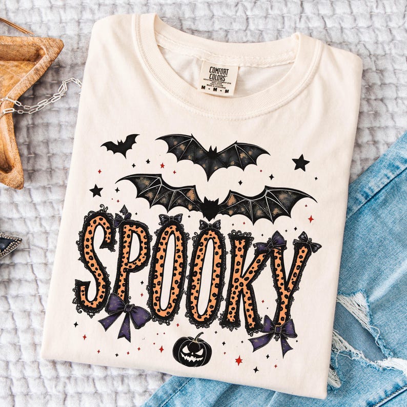 May include: Cream-colored t-shirt with a Halloween-themed design. The word "SPOOKY" is printed in orange leopard print with black lace trim and purple bows. Above the word are bats and stars, and a jack-o'-lantern is below.