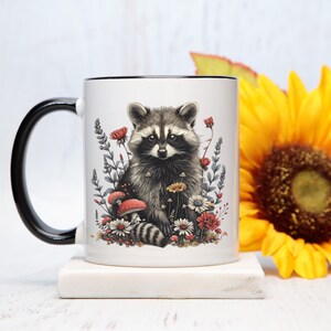 Puede incluir: Taza de cer&aacute;mica blanca con asa y borde negros, con una ilustraci&oacute;n detallada de un mapache rodeado de flores y setas. El dise&ntilde;o incluye elementos florales rojos, blancos y amarillos. La taza est&aacute; sobre una superficie blanca.
