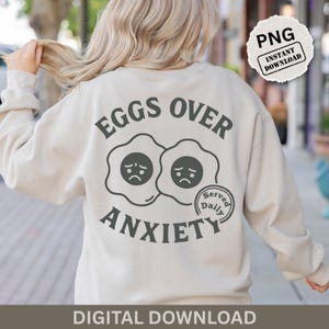 Peut inclure: Sweat-shirt crème avec l'inscription "EGGS OVER ANXIETY" en gris foncé. Le motif représente deux œufs au plat avec des visages tristes. Un petit tampon circulaire indique "Served Daily". Les mots "PNG INSTANT DOWNLOAD" sont également visibles.