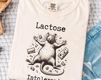 Op de afbeelding: Een beige T-shirt met een zwart-witte illustratie van een rat omringd door kaas. De tekst "Lactose Intolerant" is onder de rat gedrukt.